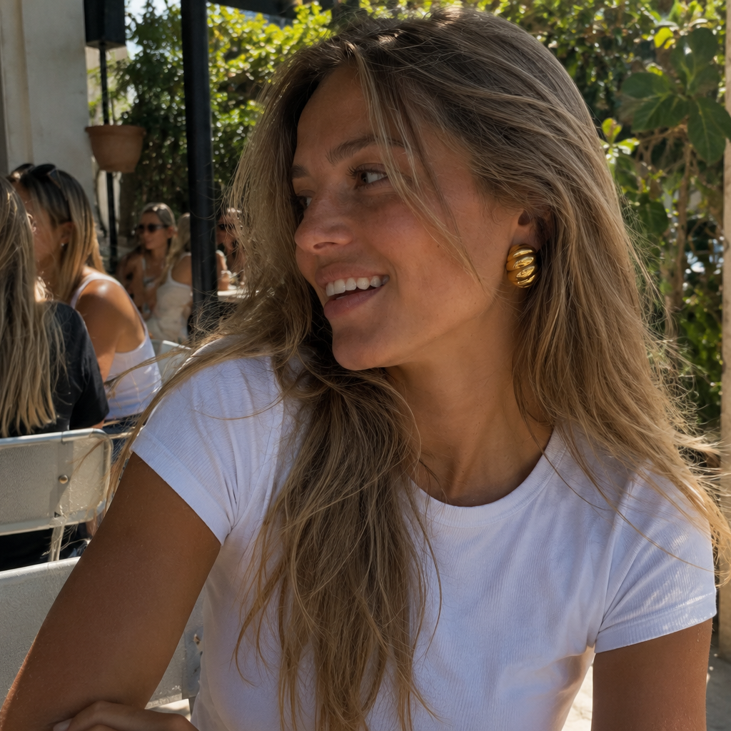 A woman with long blonde hair wearing a white t-shirt and a chunky gold hoop earring, smiling in an outdoor cafe setting.