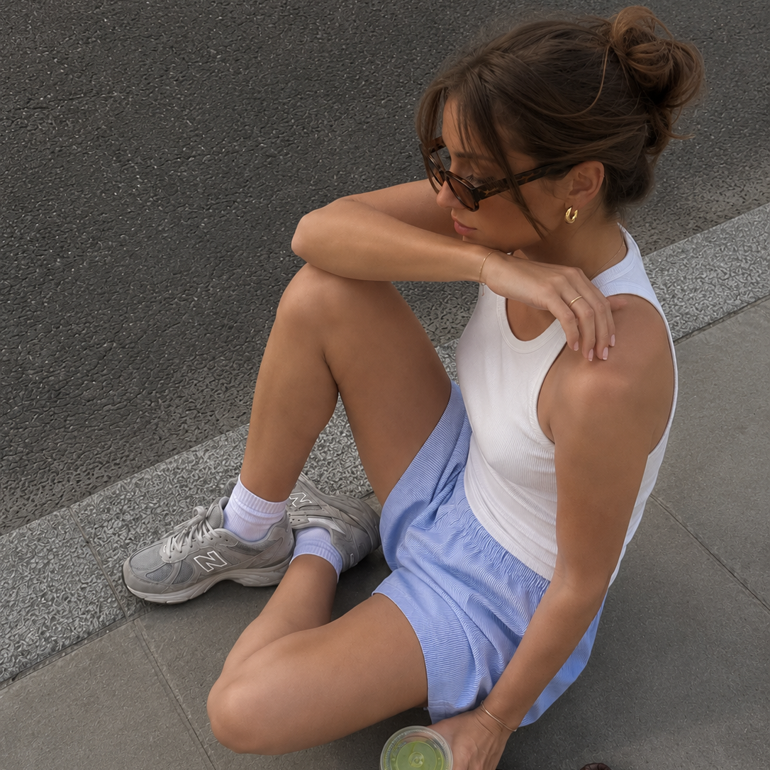Woman sitting on a sidewalk holding a green cup, wearing a white tank top and blue shorts.