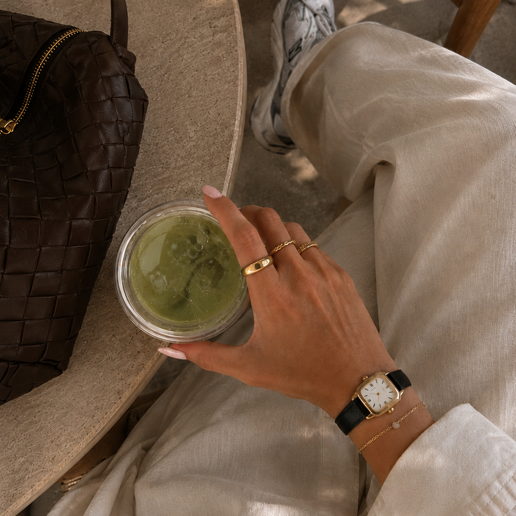 Person holding a green smoothie in a cup with a woven chair and brown bag in the background