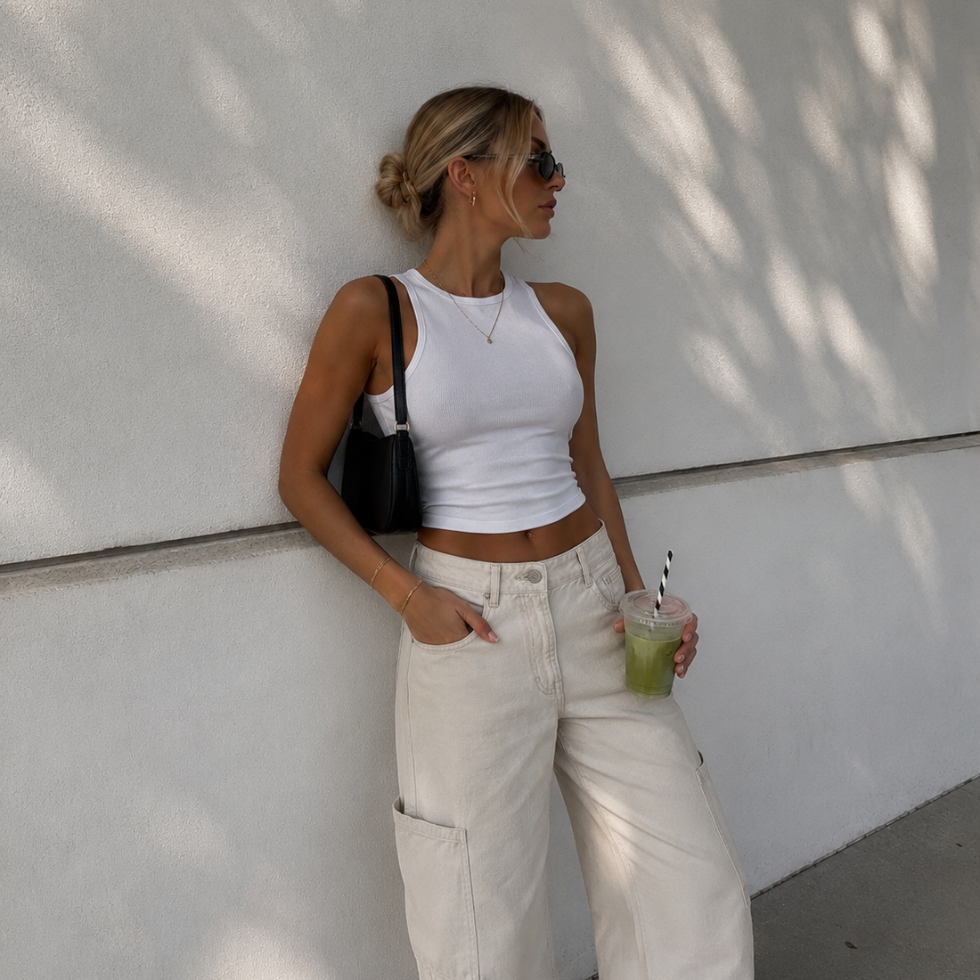 Woman in white top and beige pants holding a drink against a white wall